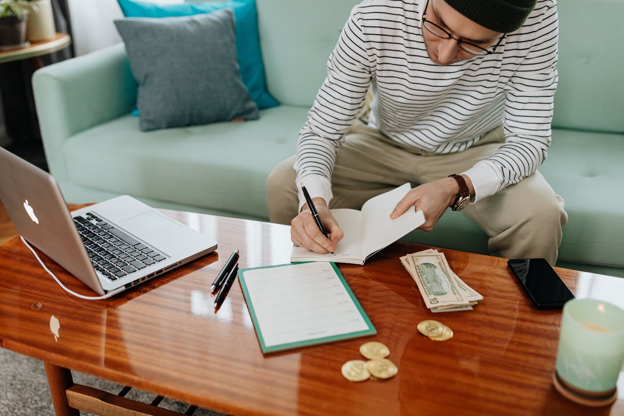 A man in a striped shirt sitting at a wooden table, writing in a notebook with money and a laptop nearby.